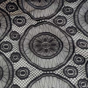 Peut inclure: Un tissu en dentelle noire pr&eacute;sentant un motif r&eacute;p&eacute;titif de grands cercles avec des centres floraux complexes, reli&eacute;s par un fond en maille hexagonale fine. Le design se compose de fils noirs denses cr&eacute;ant un textile textur&eacute; et semi-transparent.