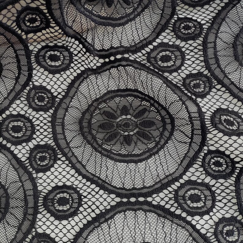 Peut inclure: Un tissu en dentelle noire pr&eacute;sentant un motif r&eacute;p&eacute;titif de grands cercles avec des centres floraux complexes, reli&eacute;s par un fond en maille hexagonale fine. Le design se compose de fils noirs denses cr&eacute;ant un textile textur&eacute; et semi-transparent.
