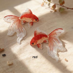 May include: Two embroidered goldfish patches featuring vibrant red and orange thread detailing on the bodies and translucent, flowing white fins with delicate red stitching. The word red is printed in small black text between the two fish.