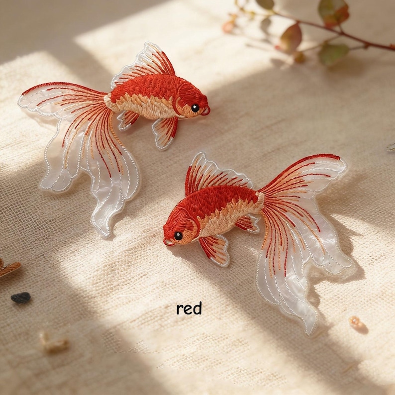 May include: Two embroidered goldfish patches featuring vibrant red and orange thread detailing on the bodies and translucent, flowing white fins with delicate red stitching. The word red is printed in small black text between the two fish.