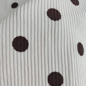 May include: A white corduroy fabric featuring a repeating pattern of dark brown polka dots. The material has a distinct vertical ribbed texture characteristic of corduroy, with the dark brown circular spots evenly distributed across the surface.