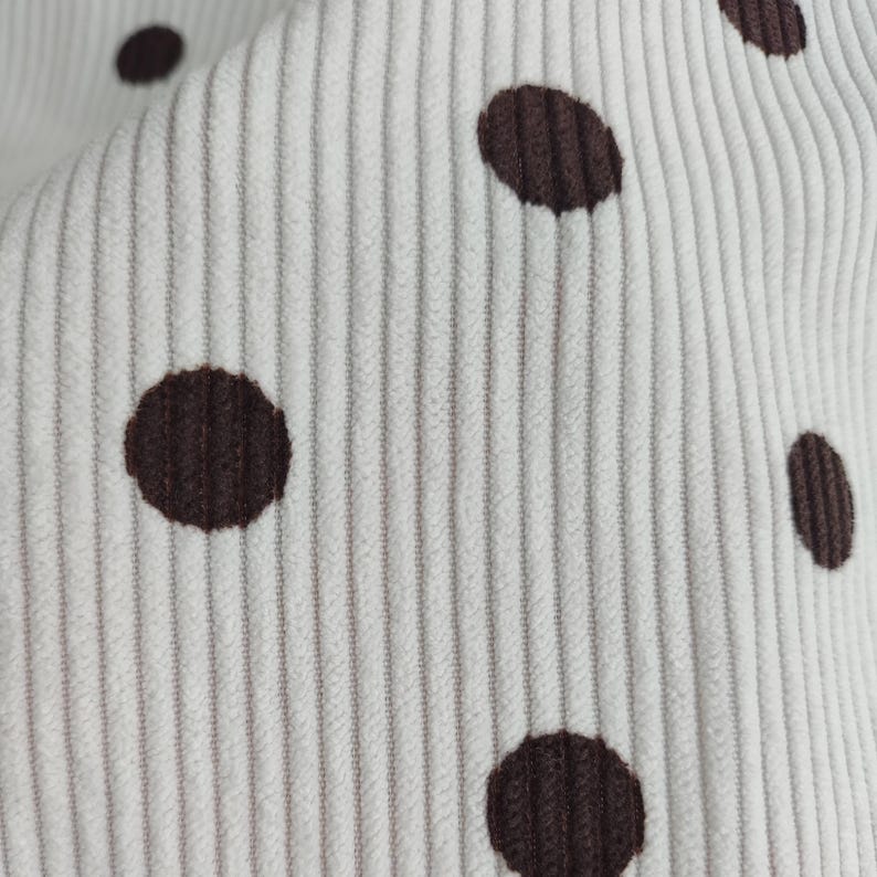 May include: A white corduroy fabric featuring a repeating pattern of dark brown polka dots. The material has a distinct vertical ribbed texture characteristic of corduroy, with the dark brown circular spots evenly distributed across the surface.