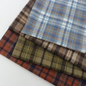 May include: A stack of four folded fabric swatches featuring various plaid patterns. The top layer is light blue and white, followed by a brown and tan layer, an olive green and dark brown layer, and a bottom layer of rust orange and dark blue.