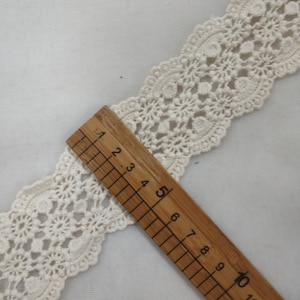 May include: A close-up of a white crocheted lace trim with a wooden ruler measuring 10 inches. The lace trim has a delicate floral pattern.