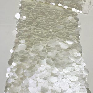 Puede incluir: Una pieza de tela de color marfil densamente cubierta con lentejuelas circulares superpuestas de varios tama&ntilde;os. El material presenta un acabado brillante y reflectante en toda la superficie, con la palabra IVORY impresa en la esquina superior izquierda.