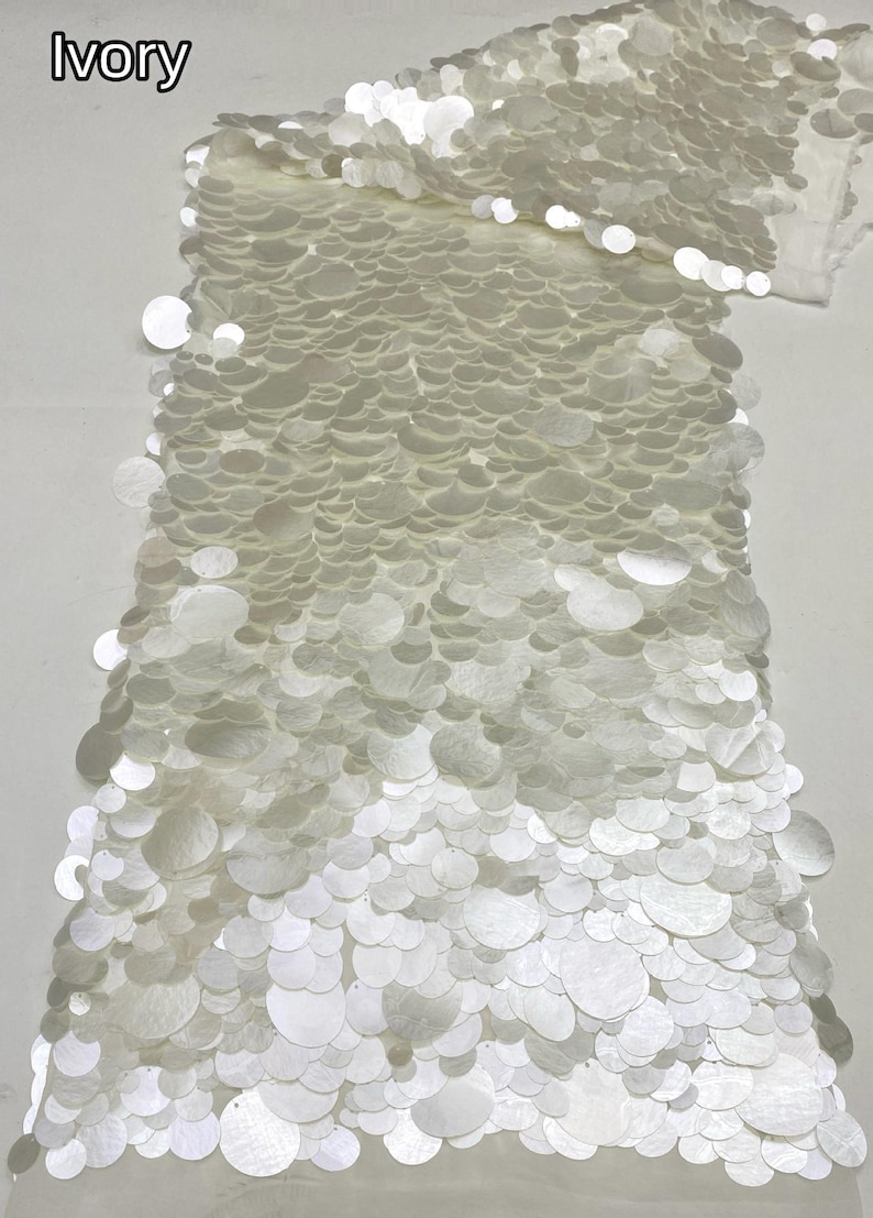 Puede incluir: Una pieza de tela de color marfil densamente cubierta con lentejuelas circulares superpuestas de varios tama&ntilde;os. El material presenta un acabado brillante y reflectante en toda la superficie, con la palabra IVORY impresa en la esquina superior izquierda.