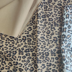 May include: Close-up of various fabrics featuring a tan solid color and leopard print patterns. The leopard prints showcase brown and black spots on a tan background, with a lighter cream-colored fabric also displaying the pattern.