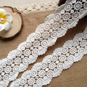 May include: White crochet lace trim with a delicate floral pattern. The lace is shown on a burlap background.