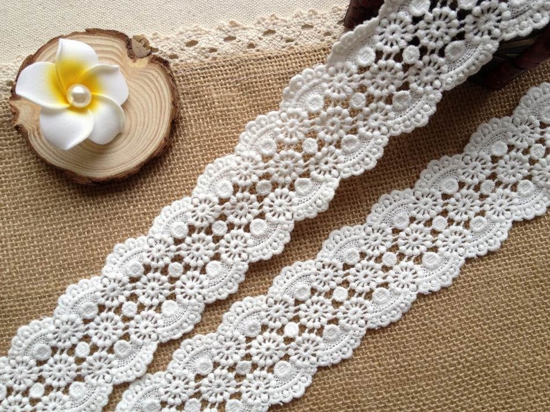 May include: White crochet lace trim with a delicate floral pattern. The lace is shown on a burlap background.