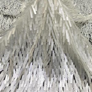 May include: A fabric textile featuring a dense layer of long, rectangular, white iridescent sequins attached to a sheer mesh base. The sequins are arranged in a vertical, overlapping pattern, creating a textured, fringe-like appearance.