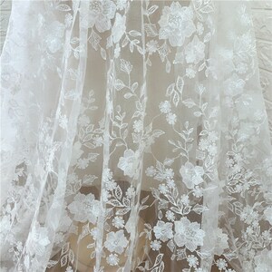 Puede incluir: Tela de encaje floral blanco con un dise&ntilde;o bordado delicado. La tela presenta patrones florales detallados con hojas y tallos, creando un aspecto texturizado y elegante. Ideal para vestidos de novia, vestidos o proyectos decorativos.
