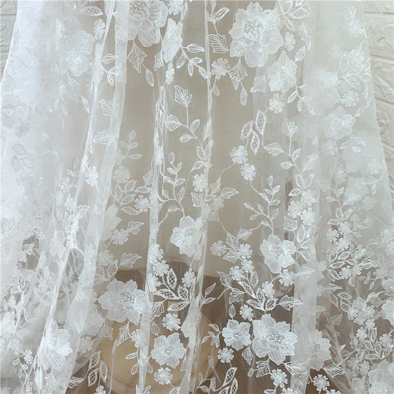 Puede incluir: Tela de encaje floral blanco con un dise&ntilde;o bordado delicado. La tela presenta patrones florales detallados con hojas y tallos, creando un aspecto texturizado y elegante. Ideal para vestidos de novia, vestidos o proyectos decorativos.