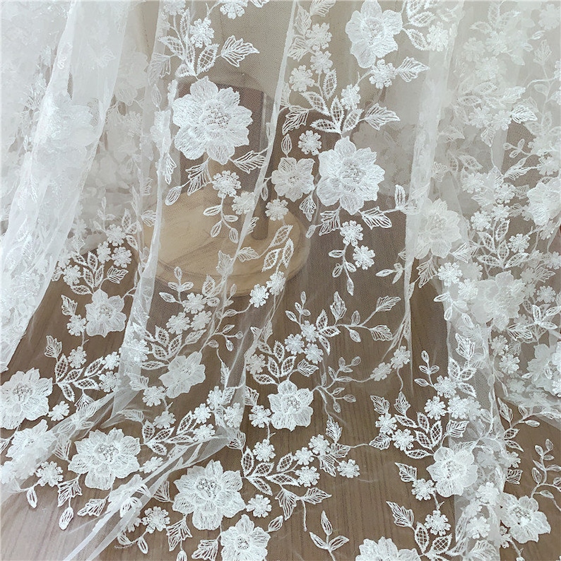 Puede incluir: Tela de encaje floral blanco con flores y hojas bordadas. El dise&ntilde;o intrincado presenta grandes flores abiertas y peque&ntilde;os acentos florales, creando una apariencia texturizada y elegante. Ideal para vestidos de novia, vestidos o proyectos decorativos.