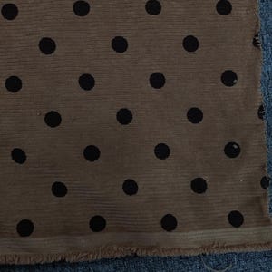 May include: A piece of tan fabric featuring a repeating pattern of solid black polka dots. The material has a visible ribbed texture and a raw, frayed edge along the bottom hem, showcasing the weave of the textile.