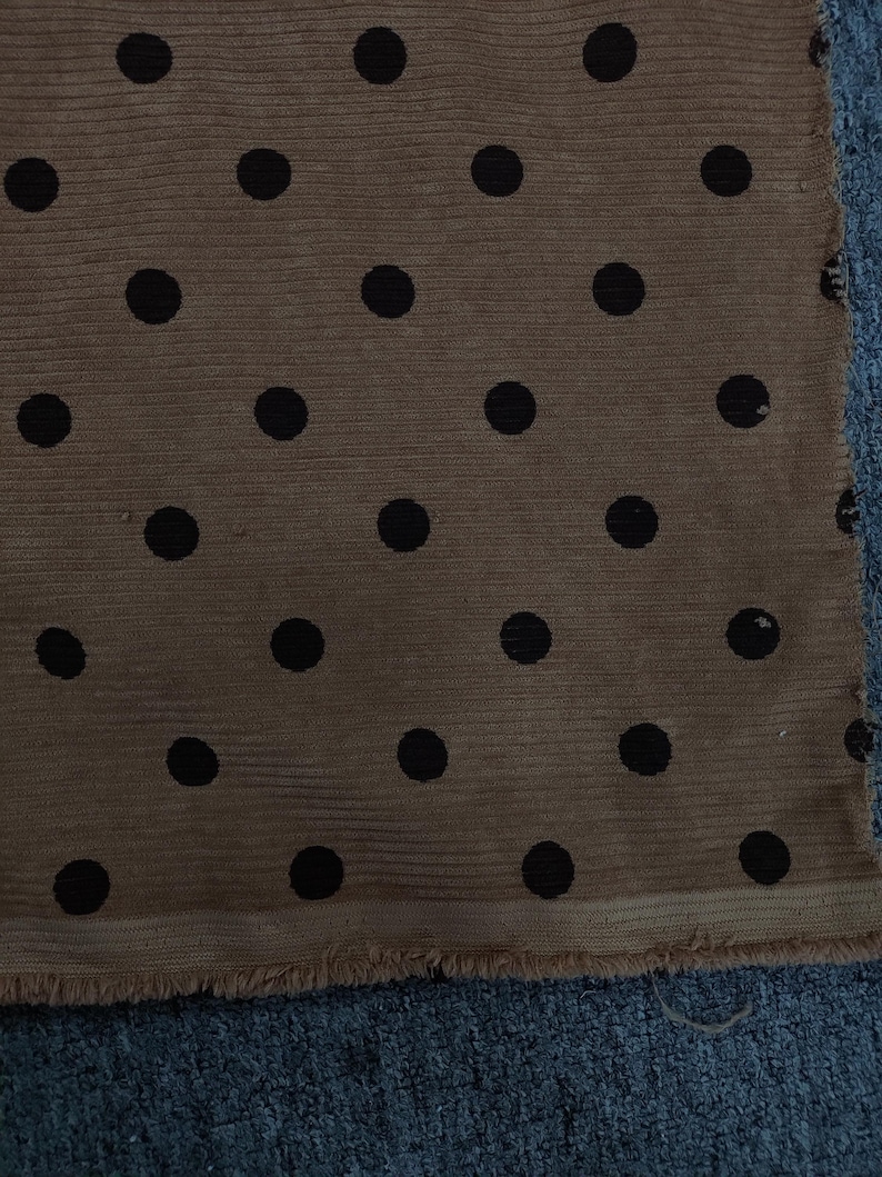 May include: A piece of tan fabric featuring a repeating pattern of solid black polka dots. The material has a visible ribbed texture and a raw, frayed edge along the bottom hem, showcasing the weave of the textile.