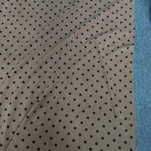 May include: A rectangular piece of fabric featuring a light tan base color with a repeating pattern of small, dark brown polka dots. The material has a visible ribbed texture and a slightly frayed edge along the bottom, showing a consistent print.