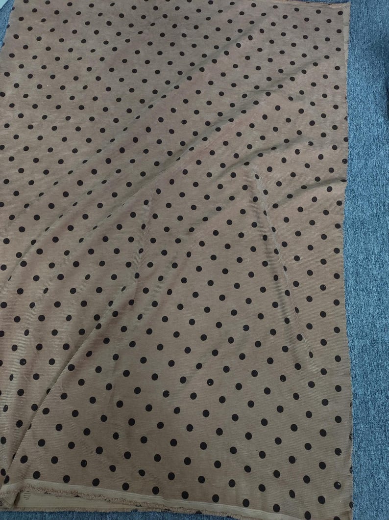 May include: A rectangular piece of fabric featuring a light tan base color with a repeating pattern of small, dark brown polka dots. The material has a visible ribbed texture and a slightly frayed edge along the bottom, showing a consistent print.