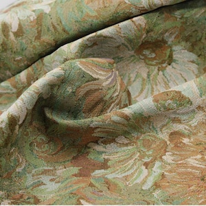 May include: A woven fabric featuring a dense floral pattern in shades of sage green, olive, and cream. The textile displays a textured, tapestry-like weave with visible interlacing threads forming large, stylized flower motifs across the surface.