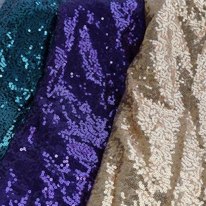 Op de afbeelding: Close-up van drie stoffen bedekt met pailletten. De stoffen zijn teal, paars en goud. De pailletten zijn dicht op elkaar gepakt, waardoor een glinsterend effect ontstaat. De stoffen zijn waarschijnlijk voor kleding of decoratieve doeleinden.