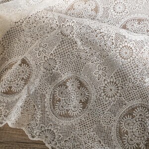 May include: Close-up of ivory lace fabric with intricate floral and geometric patterns. The delicate lace is draped, revealing the detailed design and texture. The fabric is suitable for various craft projects.