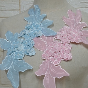 Op de afbeelding: Twee bloemvormige applicaties, een in lichtblauw en een in roze, met parelmoerachtige versieringen. De applicaties zijn gemaakt van delicate kant en hebben ingewikkelde bloemmotieven.