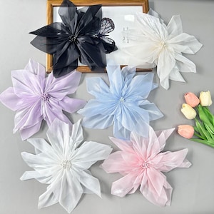 May include: A collection of six large, multi-layered fabric flowers in black, white, lavender, light blue, and pink, each featuring a central cluster of small beads. A black butterfly ornament is attached to the black flower, and a small bouquet of three peach-coloured tulip buds is positioned to the right.
