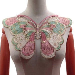 May include: A colorful butterfly applique with pink, green, and gold embroidery. The butterfly design is symmetrical, with detailed wing patterns and a delicate, openwork neckline. The applique is displayed on a mannequin.