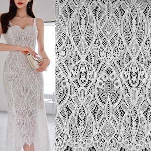 May include: A white lace dress with spaghetti straps and a fitted silhouette. The dress features an intricate floral and geometric pattern. The fabric is delicate and semi-sheer, with a scalloped hemline. A matching clutch is held in the hand.