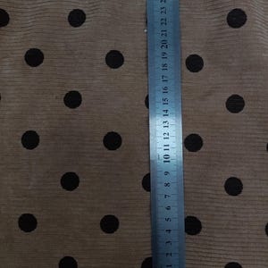 May include: A tan corduroy fabric featuring a repeating pattern of dark brown polka dots. A metal ruler is placed vertically on the fabric, showing measurements in centimeters and inches, with the ruler extending up to 28 centimeters in length.