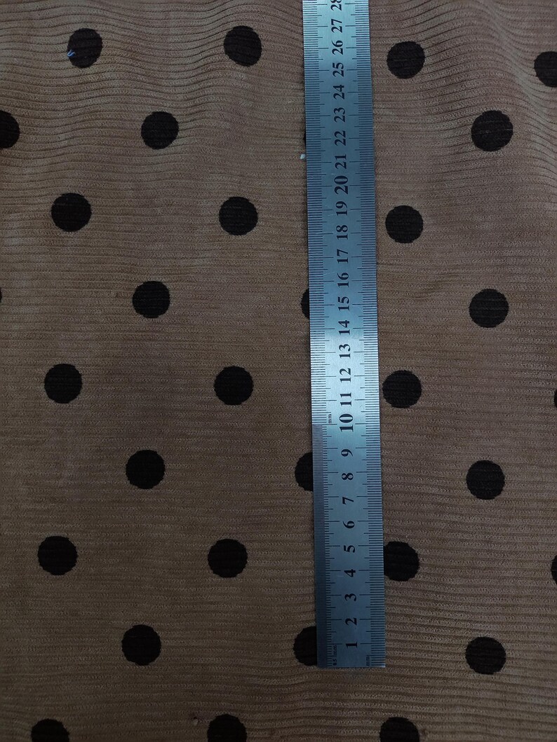 May include: A tan corduroy fabric featuring a repeating pattern of dark brown polka dots. A metal ruler is placed vertically on the fabric, showing measurements in centimeters and inches, with the ruler extending up to 28 centimeters in length.
