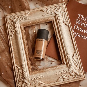 May include: A bottle of MAC Studio Fix Fluid SPF 15 foundation in a light brown shade, with the words "Studio Fix Fluid SPF 15/PA++  Skin Balancing Complex" printed on the bottle. The bottle is sitting inside a decorative, ornate, white frame.