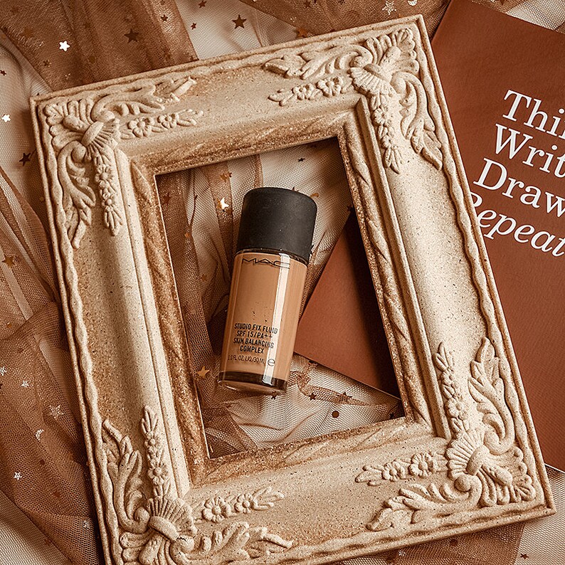 May include: A bottle of MAC Studio Fix Fluid SPF 15 foundation in a light brown shade, with the words "Studio Fix Fluid SPF 15/PA++  Skin Balancing Complex" printed on the bottle. The bottle is sitting inside a decorative, ornate, white frame.