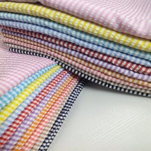 May include: A stack of colorful fabric swatches with various striped and checkered patterns. The fabrics display a range of colors, including pink, yellow, blue, red, purple, and black. The textiles are neatly arranged, showcasing the different designs and textures.