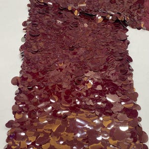 Puede incluir: Una tela de color burdeos con una capa densa de lentejuelas circulares reflectantes superpuestas. El material tiene un color rojo parduzco profundo y rico con un acabado brillante. El texto 'Burgundy' aparece impreso en la esquina superior izquierda.