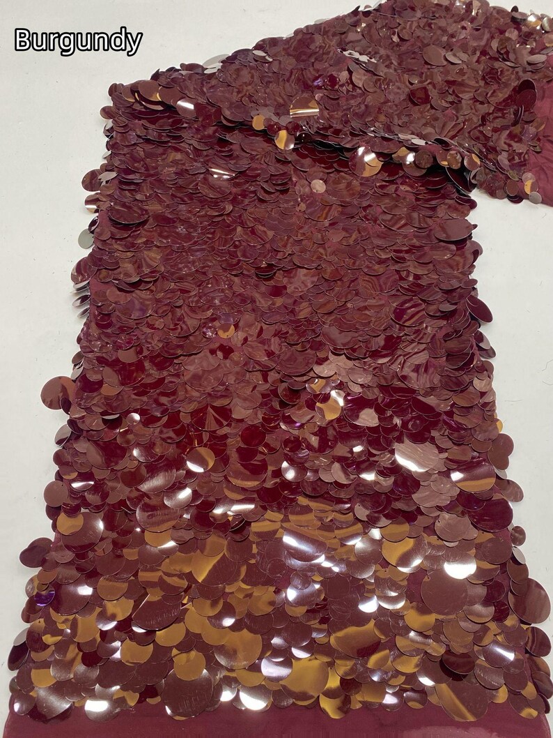 Puede incluir: Una tela de color burdeos con una capa densa de lentejuelas circulares reflectantes superpuestas. El material tiene un color rojo parduzco profundo y rico con un acabado brillante. El texto 'Burgundy' aparece impreso en la esquina superior izquierda.