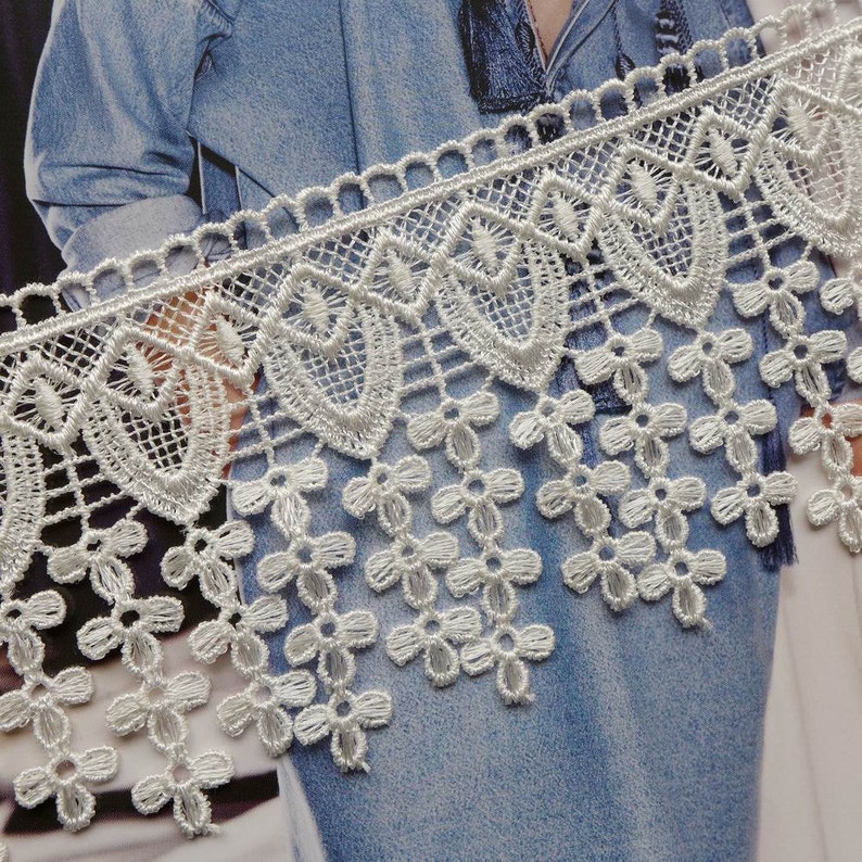 May include: White lace trim featuring a scalloped edge and a fringe of small flowers. The lace is delicate and intricate, ideal for adding a touch of elegance to any project.