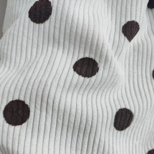 May include: A white corduroy fabric featuring a repeating pattern of dark brown polka dots. The material has a distinct vertical ribbed texture characteristic of corduroy, with the dark brown circular spots evenly distributed across the surface.