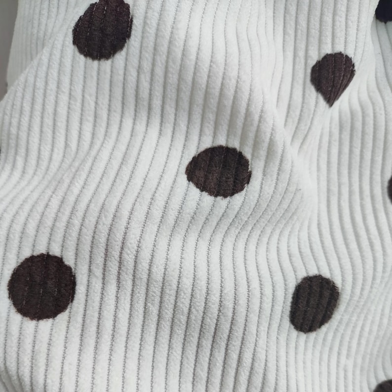 May include: A white corduroy fabric featuring a repeating pattern of dark brown polka dots. The material has a distinct vertical ribbed texture characteristic of corduroy, with the dark brown circular spots evenly distributed across the surface.