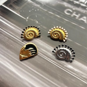 May include: Four decorative pins in gold and silver tones. Two pins are gold, spiral-shaped with black accents. One gold pin is shaped like a hand. The fourth pin is silver, spiral-shaped with black accents. The pins are on a gray surface.