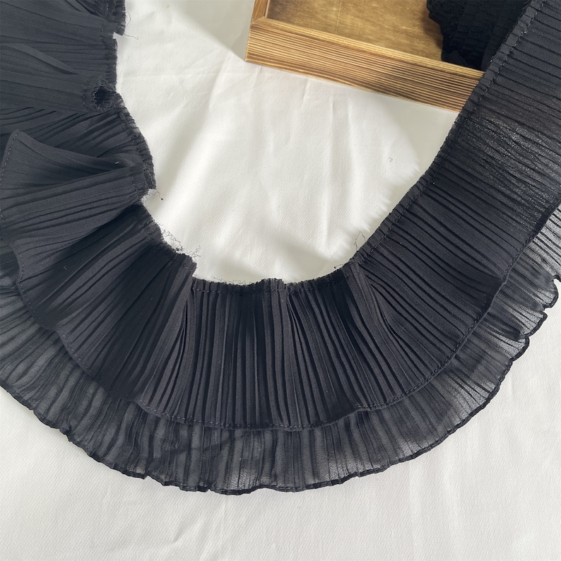 May include: Black pleated fabric trim with a layered design. The trim has a wavy, ruffled edge and is made of a sheer material. The fabric is displayed on a white surface.