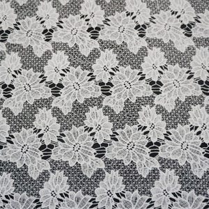 Peut inclure: Gros plan sur un tissu en dentelle florale blanche. La dentelle présente un motif répétitif de fleurs stylisées avec des motifs de pétales détaillés. Le fond est de couleur foncée, créant un contraste avec la dentelle blanche.