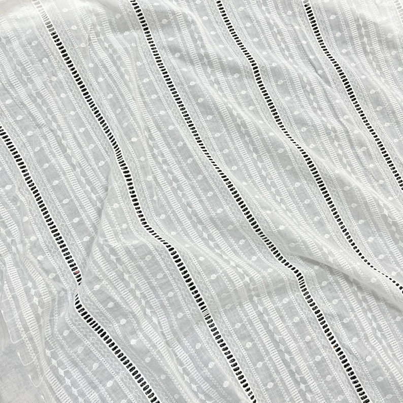 May include: White cotton fabric with a delicate embroidered pattern of small circles and lines. The fabric has a subtle sheen and a soft, drapey texture.