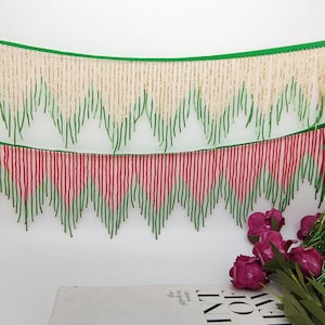 May include: Decorative trim featuring two rows of beaded fringe. The top row has gold and green beads, while the bottom row has red and green beads. The trim is against a white background, with a green border at the top.