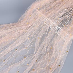 May include: A sheer, pale pink tulle fabric featuring a delicate, wavy white embroidered pattern and scattered small, round gold beads. The lightweight, translucent material is draped to showcase its fine texture and decorative embellishments.