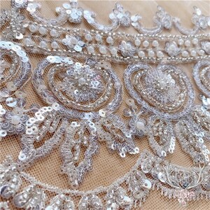 Pu&ograve; includere: Primo piano di un tessuto di pizzo bianco con perline e paillettes. Il tessuto presenta un delicato motivo floreale con perline madreperlacee e paillettes.