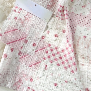 Könnte beinhalten: Ein weißer Stoff mit Patchwork-Design und verschiedenen rosa Mustern, darunter Blumenprints, Vichy-Karos und Punkte. Das Material hat eine texturierte, geknitterte Oberfläche und ist mit einem leeren, weißen, rechteckigen Papieretikett versehen.