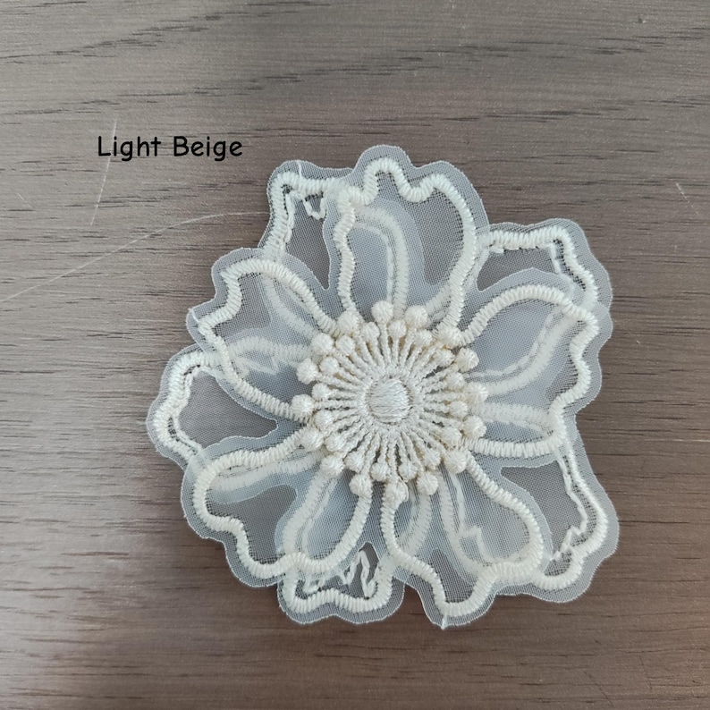 May include: A light beige flower applique featuring intricate embroidery on a sheer mesh base. The design showcases delicate scalloped petals and a textured, circular center with small raised details, labeled as Light Beige.