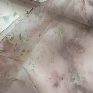May include: A sheer, lightweight fabric featuring a soft, ethereal pattern of muted mauve, dusty rose, and sage green botanical prints. The translucent material is draped in gentle folds, highlighting the delicate, watercolor-like texture.