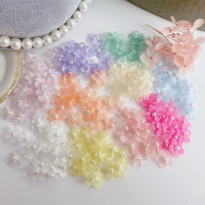 Może przedstawiać: Kolekcja kolorowych, przezroczystych ozdób z kwiatów z perłowymi środkami. Kwiaty są w odcieniach różu, fioletu, żółci, pomarańczy i błękitu. Widoczne są również suszone różowe kwiaty.