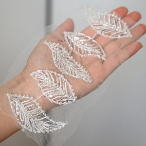 May include: Close-up of a delicate, white leaf applique. Each leaf is intricately detailed with tiny, shimmering beads and sequins, creating a sparkling effect. The leaves are attached to a sheer, white mesh fabric, perfect for embellishing bridal wear or other elegant projects.